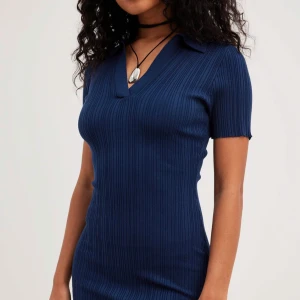 Marinblå ribbad klänning - Snygg marinblå ribbad klänning med v-ringning och krage. Klänningen har korta ärmar och en slits på sidan, vilket ger en stilren och modern look. Perfekt för både vardag och speciella tillfällen.