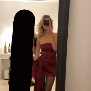 Vinröd axelbandslös klänning - Snygg vinröd axelbandslös klänning med knytning i sidan. Perfekt för en kväll ute eller en speciell tillställning. Klänningen har en kort längd och en elegant passform. Den är  omsydd än originalet till en tajtare passform