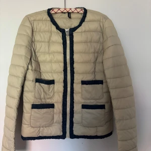 Beige quiltad jacka med svarta detaljer - Snygg beige quiltad jacka från Mango med svarta kantdetaljer och dragkedja framtill. Perfekt för en stilren look.