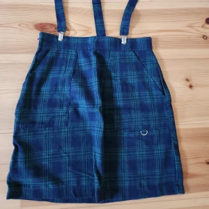Blågrön rutig kjol med hängslen - Snygg blågrön rutig kjol med justerbara hängslen och praktiska fickor. Kjolen har en D-ring detalj framtill och är perfekt för en trendig look. Passar bra till både vardag och speciella tillfällen.