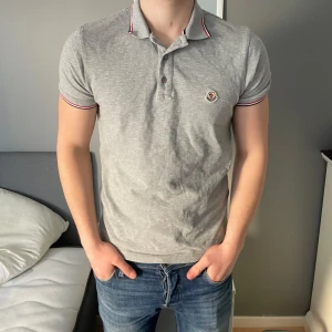 Grå pikétröja från Moncler - Säljer denna skit snygga och stilrena pikétröja från Moncler med klassisk krage och knappar framtill. Tröjan har en broderad logotyp på bröstet och röd-vit-blå detaljer på kragen. Perfekt för en avslappnad men snygg look. Mitt pris: 1099kr Nypris: 2600kr. Pris kan även diskuteras vid snabb affär. MVH, Oscar :) 