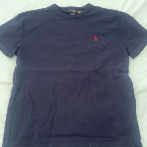 Mörkblå t-shirt från Ralph Lauren - Säljer en klassisk mörkblå t-shirt från Ralph Lauren med korta ärmar och det ikoniska röda broderade logotypen på bröstet. Perfekt för en stilren och avslappnad look.