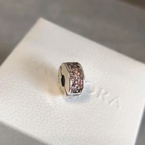 Silverberlock från Pandora - Snygg silverberlock från Pandora med glittrande rosa stenar. 💞Köpt från en annan säljare. Hon sa den är äkta men vet ej 100% men den ser exakt ut som den äkta jag köpte via butik. (kan visa konversationen med säljaren om det önskas) men tror den är äkta då kvaliten är bra!Använt den endast i två veckor ungefär men säljer för ska byta till guld clips istället. Denna har en gummi ring som följer med vilket gör att man kan sätta på den vart man vill på armbandet , antingen som avdel