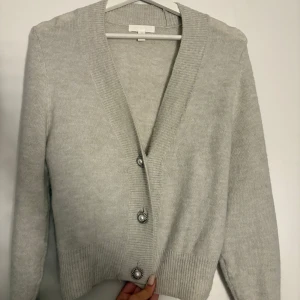 Grå kofta med pärlknappar - Säljer en elegant grå kofta med vackra pärlknappar. Koftan har en ribbad struktur och långa ärmar, perfekt för en stilren look. Den är i storlek XS och passar utmärkt för både vardag och speciella tillfällen.