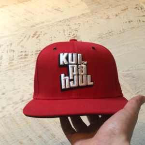 Röd keps med broderad text Kulpåhjul - Snygg röd keps med broderad text 'KUL PÅ HJUL' framtill och 'ALLTID 100%' baktill. Justerbar passform med spänne. Perfekt för en avslappnad stil.