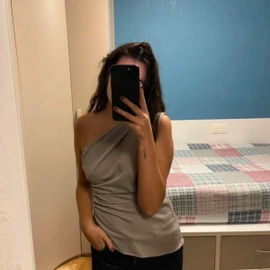 Grå oneshoulder topp - Snygg grå oneshoulder topp med draperad design. Perfekt för en stilren look. Tillverkad i ett mjukt och bekvämt material. Ny utan prislapp