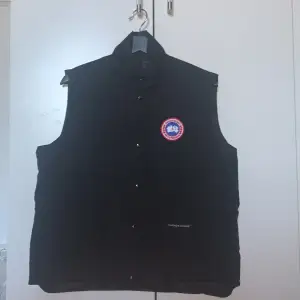 Säljer en svart dunväst från Canada Goose med deras ikoniska märke på bröstet. Västen har knappar och dragkedja framtill och är perfekt för kyligare väder. Den är tillverkad i Kanada och har hög kvalitet med Downmark-certifiering. Pris kan diskuteras, skriv om ni vill ha mer bilder