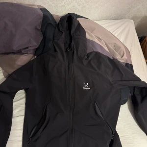 Haglöfs jacka - Säljer en stilren svart vindjacka från Haglöfs. Jackan har en praktisk huva och dragkedja framtill. Perfekt för utomhusaktiviteter och skyddar mot vind. Den har en enkel design med diskret logga på bröstet. Fraktas endast!
