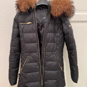 Svart dunjacka från Rockandblue - Som ny. Snygg svart dunjacka från Rockandblue med en lyxig pälsdetalj på huvan. Jackan har en quiltad design och praktiska dragkedjor i guld på fickorna och ärmsluten. Perfekt för kalla dagar.