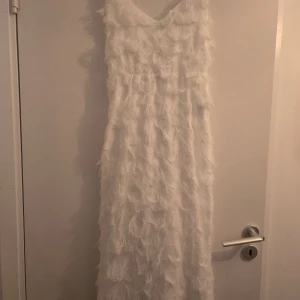 Vit klänning med fjäderdetaljer - Säljer en elegant vit klänning med fjäderdetaljer som ger en unik textur. Klänningen har en djup rygg och smala axelband, perfekt för speciella tillfällen. Den är lång och har en v-ringad halslinje. Perfekt för dig som vill sticka ut!