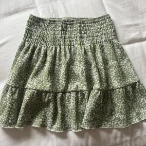 Grön blommig volangkjol Gina Tricot - Supersöt grön kortkjol från Gina Tricot med vitt blommönster och volanger. Kjolen har smock i midjan för en bekväm passform och är perfekt för varma dagar. ☺️