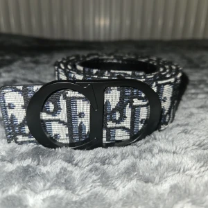 Svartvit Dior bälte med monogram - Snyggt bälte från Dior med klassiskt svart och vitt monogrammönster och stor CD-spänne i matt svart metall. Bältet är tillverkat i textil med detaljer i skinn vid hålen. Perfekt accessoar för att lyfta din outfit med en lyxig touch.