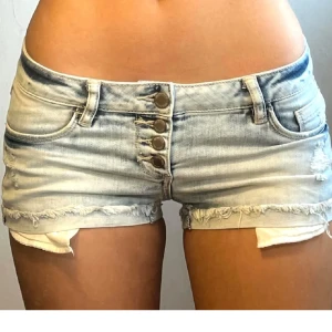 Lågmidjade ljusa jeansshorts - Snygga lågmidjade jeansshorts i ljusblå denim med slitna detaljer och fransig kant. Shortsen har flera knappar framtill, klassiska fickor både fram och bak samt en tight passform. Perfekta för varma sommardagar och en avslappnad look. Shortsen är lågmidjade!