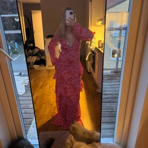 Rosa blommig långklänning med volanger - Säljer en superfin långklänning i rosa med blommigt mönster. Klänningen har djup v-ringning, långa ärmar och flera lager volanger nedtill som ger en romantisk vibe. Perfekt för dig som vill sticka ut med en färgglad och flowy look.