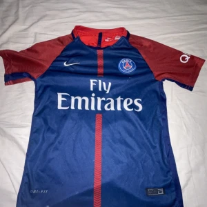 PSG Neymar Jr matchtröja Nike blå/röd - Paris Saint-Germain fotbollströja med Neymar Jr och nummer 10 på ryggen. Tröjan är blå med röda ärmar och detaljer, har Fly Emirates-tryck på bröstet och är tillverkad i Dri-FIT-material. Klassisk Nike-logga och PSG-märke på framsidan. Kortärmad modell.