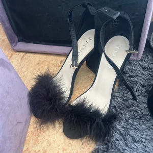 Svarta sandaler med päls från Tiamo - Svarta sandaler från Tiamo med blockklack och öppen tå. Remmar runt vristen med spänne och fluffig pälsdetalj framtill. Skorna är i mockaliknande material och har en trendig, glamorös vibe som sticker ut.