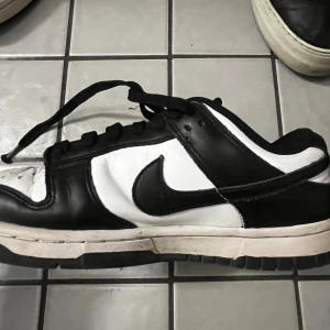 Nike Dunk Low svart/vit sneakers - Nike Dunk Low sneakers i svart och vitt skinn med klassisk Swoosh-logga på sidorna. Lågt skaft, svart snörning och perforerad tåbox för extra ventilation. Perfekt för dig som gillar streetstyle och vill ha en ikonisk look.