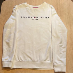 Vit sweatshirt från Tommy Hilfiger - Vit sweatshirt från Tommy Hilfiger med broderad logga och text 'EST. 1985' på bröstet. Det är till för barn/ungdom i storlek 152. Den är inprincip oanvänd. Klassisk rund halsringning med ribbade muddar och diskreta Hilfiger-detaljer. Perfekt för en clean och stilren look. 