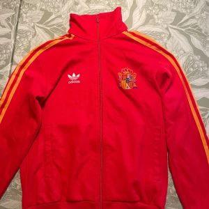 Retro Adidas track jacket Spanien S - Röd Adidas track jacket med Spaniens landslagsemblem broderat på bröstet. Klassiska gula ränder längs ärmarna, hög krage och hel dragkedja framtill. Perfekt för dig som gillar sportig stil och vill sticka ut med färg. Vintage tröja men otroligt välbevarad för sin ålder