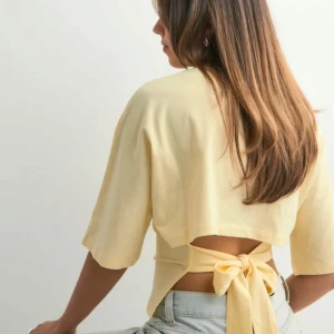 Gul omlottopp med knyt i ryggen - Snygg ljusgul topp med omlottdesign och stor knytrosett i ryggen. Toppen har halvlånga ärmar och croppad passform som visar lite rygg. Perfekt för dig som vill ha en trendig och luftig look till sommaren.