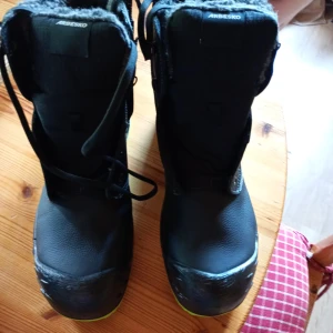 Arbesko svarta skyddskängor stl 46 - Svarta skyddskängor från Arbesko med gröna sulor och snörning. Fodrade med mjukt material för extra värme och komfort. Tillverkade i slitstarkt skinn och syntet, perfekta för kalla dagar och tuffa miljöer. Snygg detalj med svensk flagga på sidan.