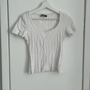 Vit ribbad topp från BikBok - Supersöt vit ribbad topp från BikBok med korta ärmar och rundad urringning. Toppen har diskreta vertikala ränder och en liten rosett framtill som detalj. Perfekt att styla med jeans eller kjol för en fräsch look.