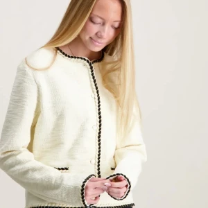 Vit cardigan jacka med svarta detaljer - Snygg vit cardigan jacka med svarta tvinnade kanter och pärlknappar. Jackan har rund halsringning, två små fickor framtill och är långärmad. Perfekt för dig som gillar en klassisk men trendig look.