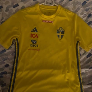 Svenska landslagets tröja Adidas - Gul fotbollströja från Adidas med svenska landslagets emblem på bröstet. Tröjan har blå detaljer, klassiska Adidas-ränder på ärmarna och flera sponsorer tryckta på framsidan. Tillverkad i lätt och ventilerande material, perfekt för träning eller match.