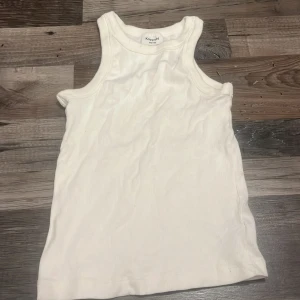 Vit ribbad linnetopp från Kappahl - En klassisk vit ribbad linnetopp från Kappahl i storlek 146/152. Toppen har ärmlös design med breda axelband och rund halsringning. Perfekt för varma dagar eller som lager under andra plagg. Enkel och stilren look som passar till det mesta.