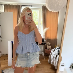 Blåvit randig linnetopp med knappar - Supersöt blå och vit randig linnetopp med smala axelband och knappar framtill. Toppen har en figurnära passform upptill och en utsvängd, peplum-liknande nederdel som ger en feminin siluett. Perfekt för varma dagar och enkel att styla med jeans eller kjol.