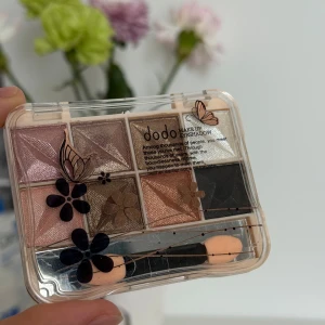 Dodo ögonskuggspalett med 8 nyanser - Palett med 8 skimrande ögonskuggor i rosa, brunt, beige, svart och champagne. Kommer i en genomskinlig ask med blommor och fjärilar på locket. Inkluderar två små applikatorer. Perfekt för att skapa både naturliga och dramatiska looks.