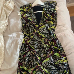 Glittrig paljettklänning från Zara - Säljer en kort, glittrig klänning från Zara med djup v-ringning. Klänningen har ett färgstarkt paljettmönster i svart, silver, grönt och lila. Perfekt för dig som vill sticka ut med en unik look. Materialet är syntet och klänningen är ärmlös.