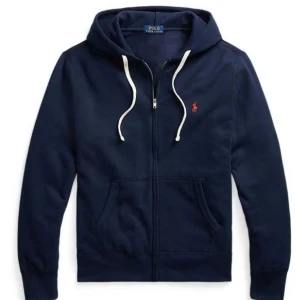 Marinblå hoodie från Polo Ralph Lauren - Snygg marinblå hoodie från Polo Ralph Lauren med dragkedja, huva och vita snören. Klassisk röd logga broderad på bröstet och känguruficka framtill. Perfekt för dig som gillar stilren och sportig look.