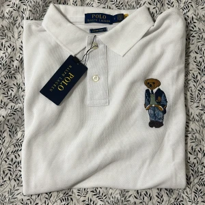 Vit pikétröja Polo Ralph Lauren björn - Vit pikétröja från Polo Ralph Lauren med klassisk krage och knäppning framtill. På bröstet finns en broderad björn i jeans och kavaj. Tröjan är i bomull och har korta ärmar. Perfekt för en clean och stilren look.