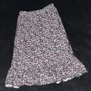 Blommig volangkjol i svart och rosa - Supersöt svart kjol med små rosa och vita blommor, volangkant nertill och resår i midjan. Perfekt för dig som gillar romantisk och lekfull stil. Lätt och skön att bära, passar till sneakers eller sandaler.