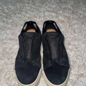 Svarta slip-on sneakers från Eytys - Svarta slip-on sneakers från Eytys i mocka med vit platt sula och lila band bak på hälen. Skorna har minimalistisk design, rund tå och diskret logga på häl och plös. Insidan har korksula och detaljer i svart skinn. Perfekta för en clean streetstil.