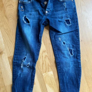Philipp Plein blå slitna jeans - Blå jeans från Philipp Plein med straight cut, coola slitningar och trasiga detaljer framtill. Snygga silverfärgade knappar och dödskalle-detaljer vid fickan. Klassisk femficksmodell med röd söm på insidan av linningen. Perfekta för en edgy streetstil.
