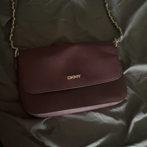 Vinröd axelväska från DKNY i skinn - Snygg vinröd axelväska från DKNY i skinn med guldfärgad kedjerem och logga framtill. Väskan har ett stilrent, fyrkantigt format och innerfoder med DKNY-mönster samt dragkedjefack. Perfekt för dig som vill ha en trendig och praktisk väska.