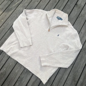Ljusgrå half zip tröja Polo Ralph Lauren - Ljusgrå långärmad tröja från Polo Ralph Lauren med klassisk half zip-dragkedja och broderad logga på bröstet. Mjuk bomullskvalitet och ribbade muddar vid ärmslut och krage. Perfekt för en clean och avslappnad stil.