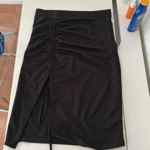 Svart kjol med snörning och slits - Svart kjol i midi-längd med snygg snörning och rynkdetalj framtill. Kjolen har en slits på ena sidan som ger en cool vibe och är tillverkad i ett mjukt, stretchigt material. Perfekt för dig som gillar stilrena plagg med en twist.