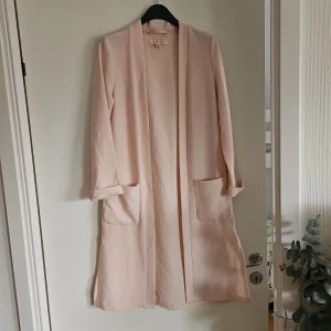 Ljusrosa kappa. - Säljer en stilren ljusrosa kappa från med raka linjer och stora fickor framtill. Jackan har lång ärm och öppen front utan knappar eller dragkedja. Perfekt för dig som gillar minimalistisk och clean stil.