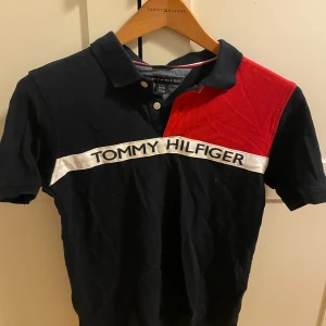 Tommy Hilfiger pikétröja svart/röd - Snygg pikétröja från Tommy Hilfiger i svart med röd axel och vit logga över bröstet. Klassisk krage, korta ärmar och två knappar framtill. Perfekt för dig som gillar sportig och stilren look. Materialet är mjuk bomull.