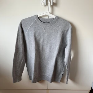 Grå stickad tröja från Superdry Studios - Snygg grå stickad tröja från Superdry Studios i storlek M. Tröjan har rund halsringning, ribbade muddar och lång ärm. Enkel och stilren design som passar perfekt till jeans eller chinos. Materialet är mjukt och skönt mot huden.
