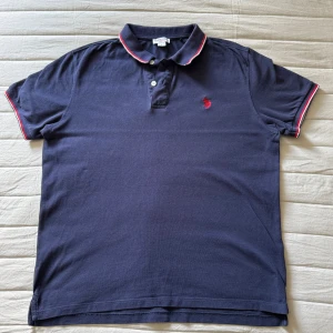 Mörkblå pikétröja från U.S. Polo Assn. - Snygg mörkblå pikétröja från U.S. Polo Assn. Röd broderad logga på bröstet och vita/röda detaljer på krage och ärmslut. Perfekt nu till sommaren 