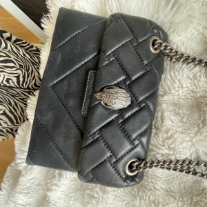 Svart quiltad axelväska från Kurt Geiger - Snygg svart quiltad axelväska från Kurt Geiger med silverfärgad kedjerem och ikoniskt örndekor på locket. Väskan är tillverkad i skinn och har ett lyxigt mönstrat innerfoder. Köpt i London på deras butik i Harrods. Köpte då med euro för cirka 2 år sedan men översatt var nypriset kring 3500kr. Lite tecken på användning men inte sliten. Skulle nog kunna hitta dustbag och annat äktehetsbevis vid önskan🥰