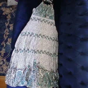 Boho långklänning med paisleymönster - Snygg långklänning i boho-stil med paisley- och blommönster i turkos, brun och beige. Klänningen har halterneck med knytband och dekorativa träknappar samt ringar vid axlarna. Luftigt material, perfekt för varma dagar och festivalvibbar.