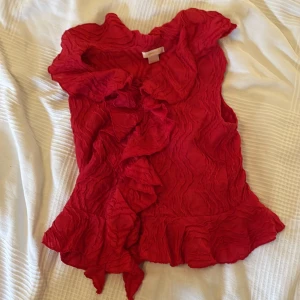 Röd volangblus från Gina Tricot - Supersöt röd blus från Gina Tricot med volanger längs halsringningen och framtill. Blusen har en crinklad struktur och är ärmlös, perfekt för att sticka ut med en färgstark look. Passar dig som gillar detaljer och vill ha något unikt i garderoben.