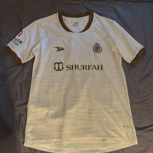 Al Nassr Ronaldo 7 fotbollströja vit/guld - Snygg Al Nassr fotbollströja med Ronaldo 7 på ryggen. Tröjan är vit med guldiga detaljer, bruna muddar och krage. Tillverkad i lätt och ventilerande material, perfekt för match eller träning. Tryckta loggor och sponsortryck på bröst och ärm.