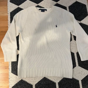 Vit stickad tröja från Ralph Lauren - Klassisk vit stickad tröja från Ralph Lauren i 100% bomull. Tröjan har rund halsringning, ribbade muddar och nederkant samt den ikoniska broderade loggan på bröstet. Perfekt för en clean och stilren look.