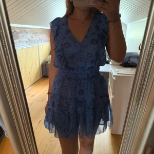 Blå blommig volangklänning - Supersöt blå kortklänning med blommigt mönster och volanger. Ett litet hål men det är inget som syns när klänningen är på